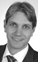 Porträt eines Mannes mit kurzem, gefärbtem Haar, der in einem Anzug und einer Krawatte auf einem neutralen Hintergrund lächelt. Er zeigt eine freundliche Ausstrahlung und präsentiert sich professionell.